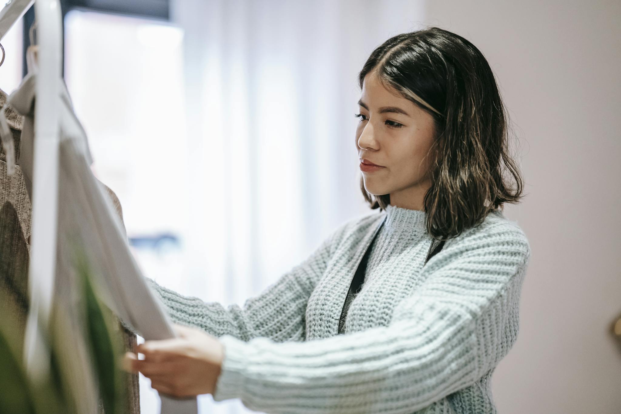A young woman in a cozy sweater browsing clothes indoors with a thoughtful expression.