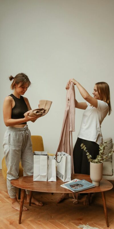 Two young women at home enjoying their new shopping purchases, displaying clothes and gift bags.